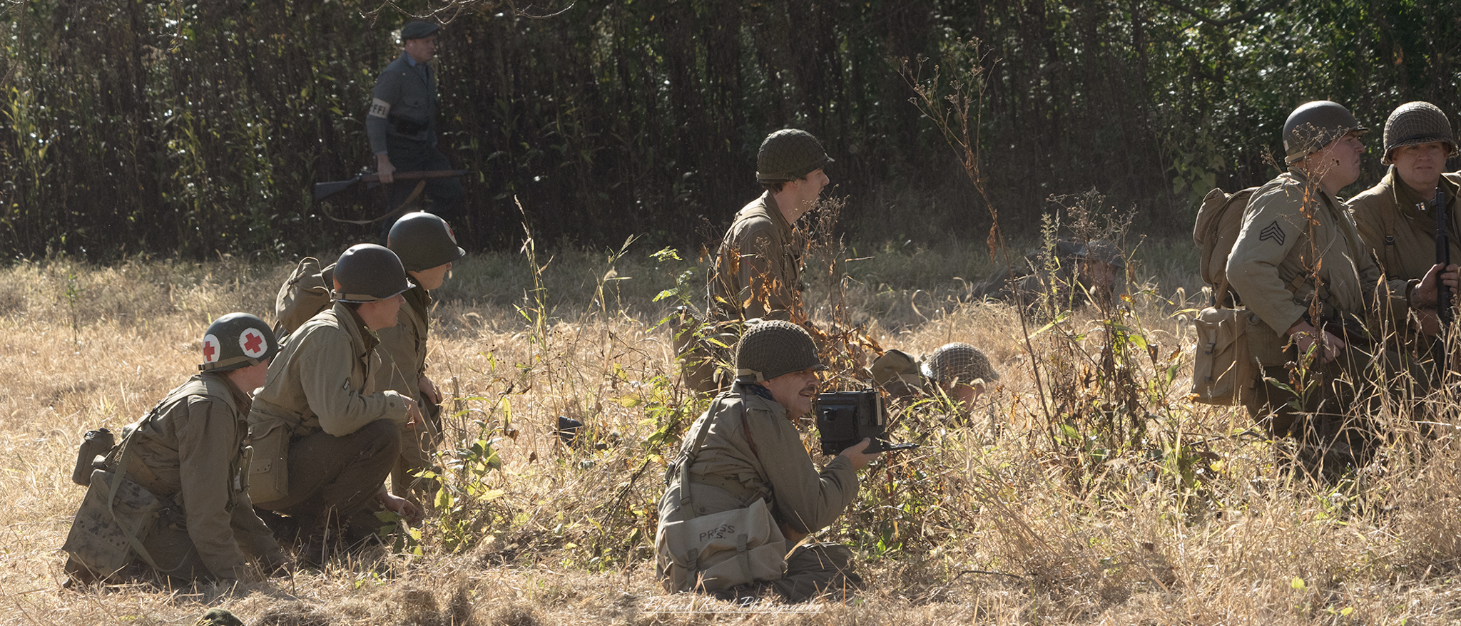 "A combat photographer in the field, crouching low with a camera in hand, capturing the intense scenes of battle. Dressed in military gear with a helmet and combat vest, the photographer blends into the rugged landscape, documenting the realities of war amidst smoke and debris. The photographer's focus is unwavering, portraying the critical role of those who risk their lives to capture history in the making."